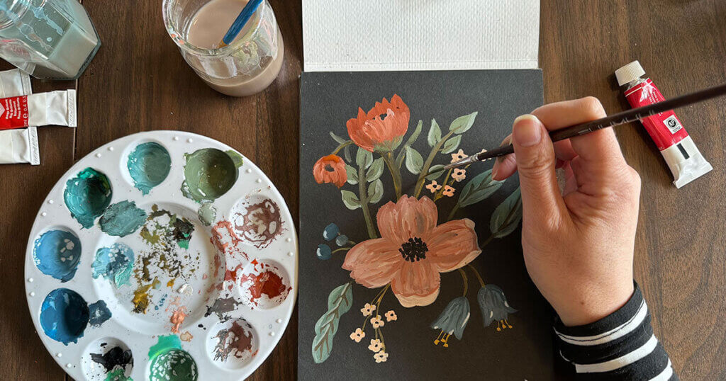 A person paints botanical flowers with gouache on black paper using a brush, surrounded by a palette of paints, tubes, and a cup of water on a wooden table.